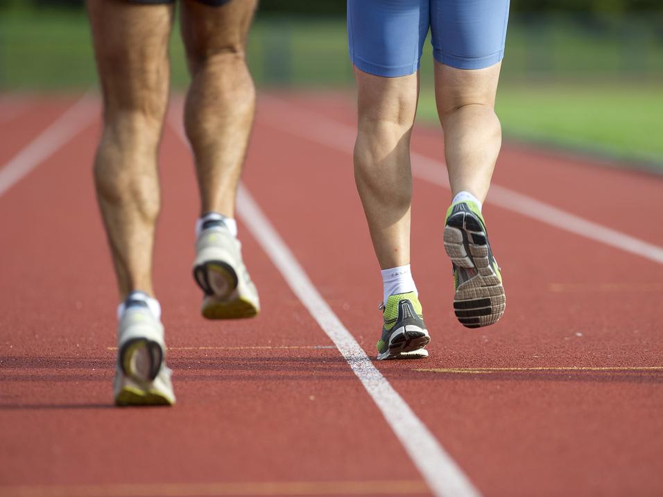 Zwei Läufer, von hinten gesehen, joggen auf einer roten Laufbahn. Der eine trägt kurze Sportshorts, der andere eine blaue Hose.