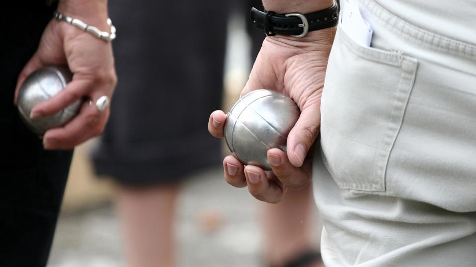 Zwei Personen halten silberne Kugeln in der Hand, während sie auf einem Platz an einem Boule-Spiel teilnehmen.