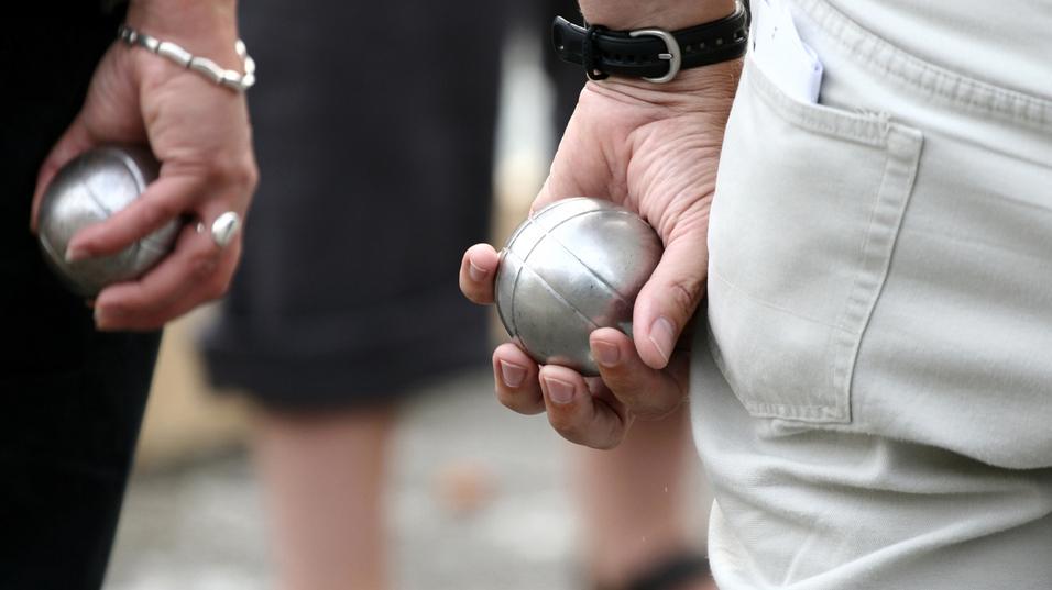 Zwei Personen halten silberne Kugeln in der Hand, während sie auf einem Platz an einem Boule-Spiel teilnehmen.