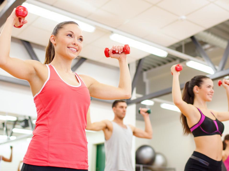 Vier Personen heben Gewichte in einem Fitnessstudio. Zwei Frauen tragen Sporttops, ein Mann hat ein T-Shirt an.