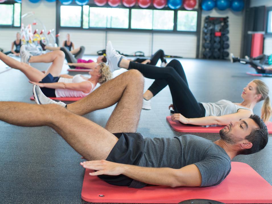 Gruppe von Personen macht Übungen auf Fitnessmatten in einem modernen Gym mit Fitnessgeräten im Hintergrund.