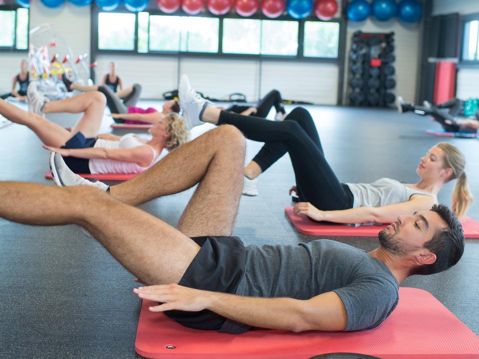 Gruppe von Personen macht Übungen auf Fitnessmatten in einem modernen Gym mit Fitnessgeräten im Hintergrund.