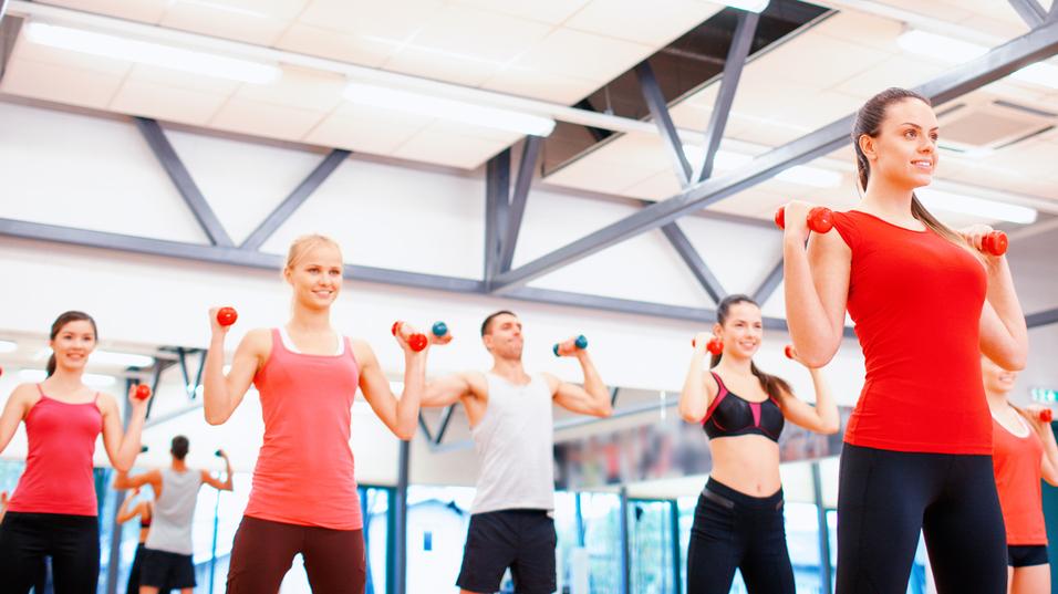 Gruppe von Menschen in Sportkleidung hebt Hanteln in einem Fitnessstudio mit Spiegeln und heller Beleuchtung.