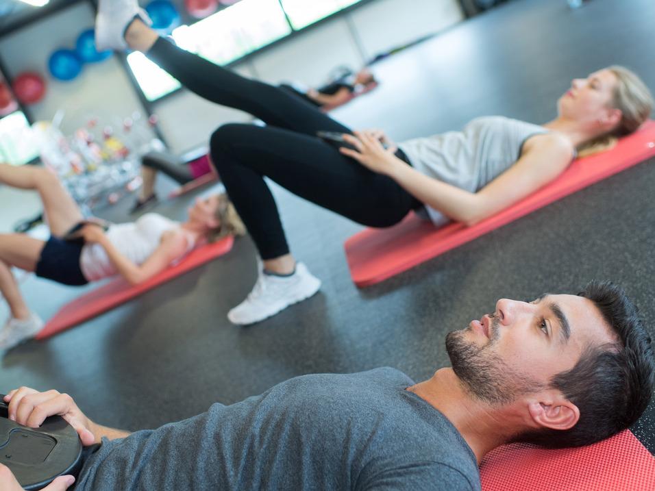Gruppe von Menschen bei einem Workout, einige auf Fitnessmatten mit Gewichten, andere in Bauchlage.