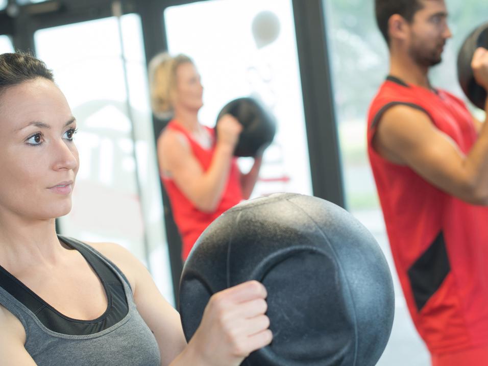 Drei Personen im Fitnessstudio trainieren mit schweren Bällen; eine Frau steht im Vordergrund mit fokussiertem Blick.