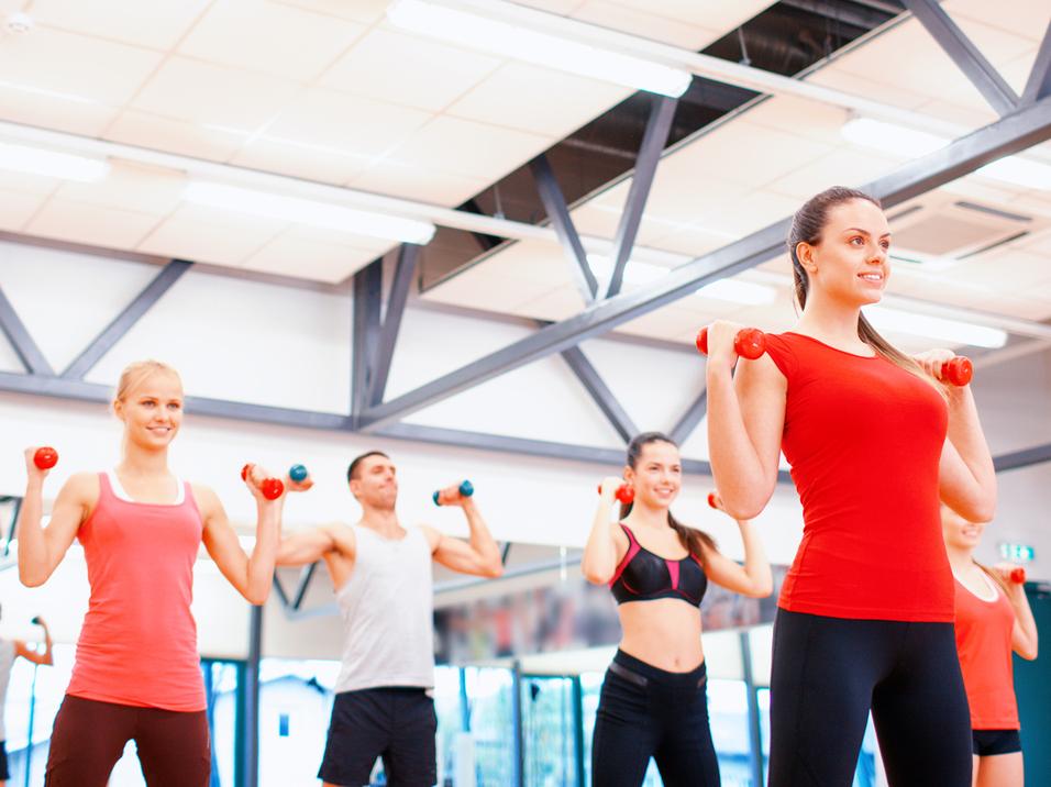 Gruppe von Menschen in Sportkleidung hebt Hanteln in einem Fitnessstudio mit Spiegeln und heller Beleuchtung.