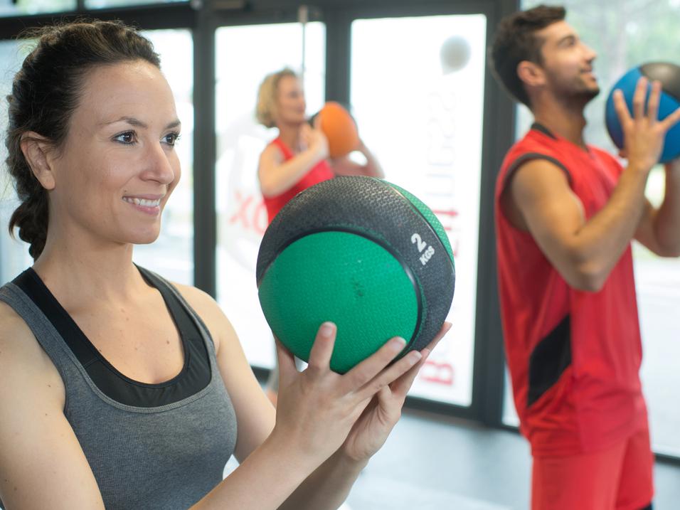 Drei Personen trainieren im Fitnessstudio mit Trainingsbällen; eine Frau hält einen grünen Medizinball.