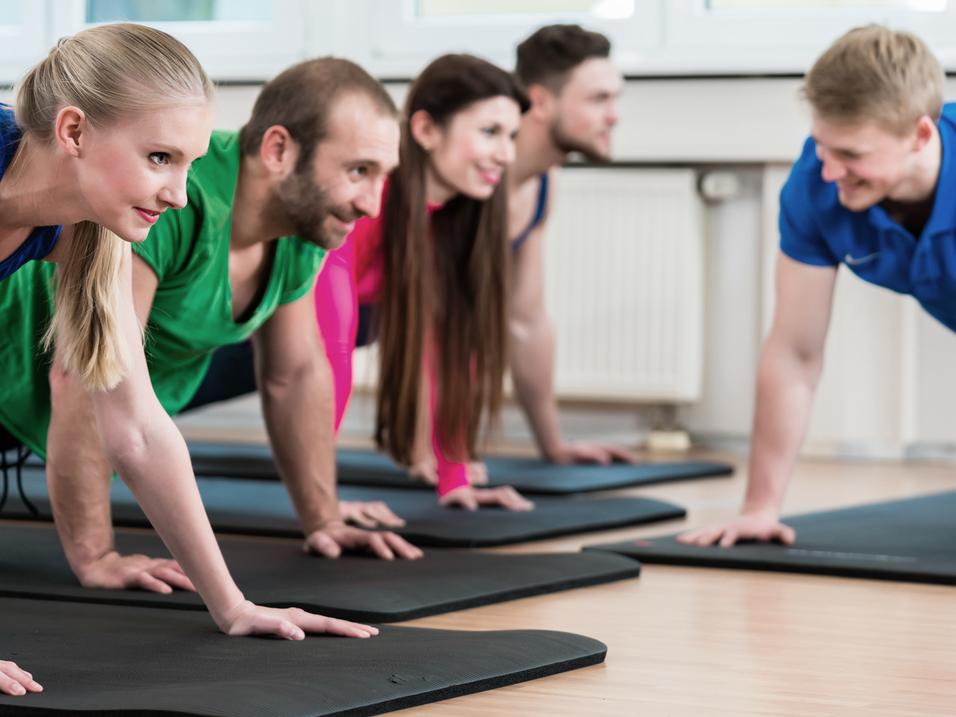 Fünf Personen in Sportbekleidung machen Liegestütze auf Matten in einem hellen Fitnessraum.
