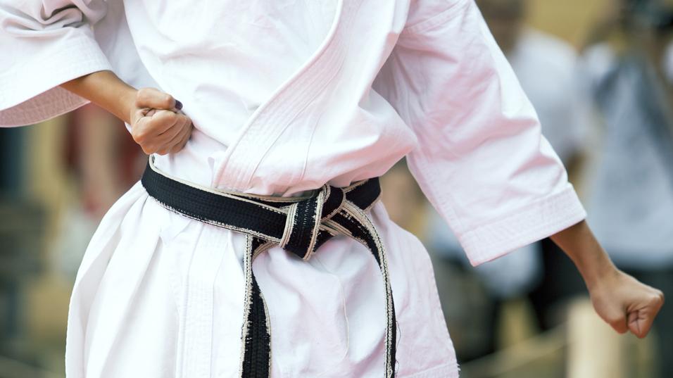 Nahaufnahme eines Karateka in Weiß mit schwarzem Gürtel, der eine Faustballenposition einnimmt.