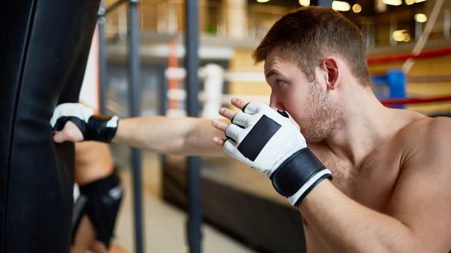 Boxer schlägt mit einem Handschuh auf einen Trainingsdummy, im Hintergrund weitere Boxsport-Aktivitäten.