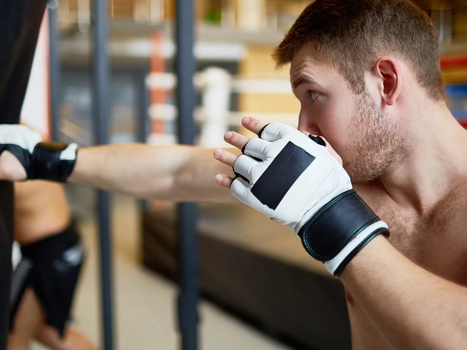 Boxer schlägt mit einem Handschuh auf einen Trainingsdummy, im Hintergrund weitere Boxsport-Aktivitäten.