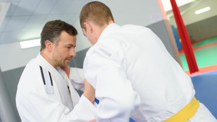 Zwei Judoka in weißen Kimonos üben eine Technik im Training auf einer blauen Matte. Ein Judoka trägt einen gelben Gürtel.