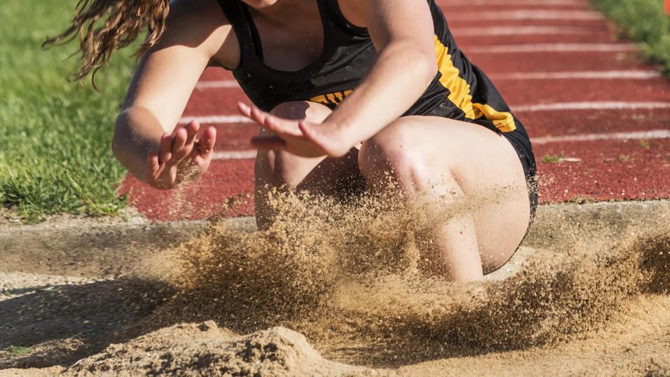 Athletin springt in einen Sandgruben, während Sand aufwirbelt. Grüße des Sprungbretts sind im Hintergrund sichtbar.
