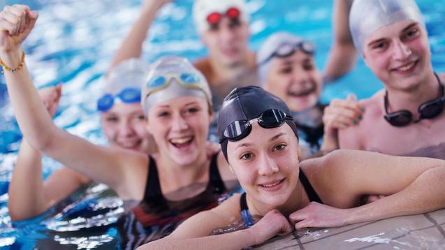 Sechs Jugendliche in Badekappen und Schwimmbrillen posieren fröhlich im Wasser, einige mit erhobenen Armen.