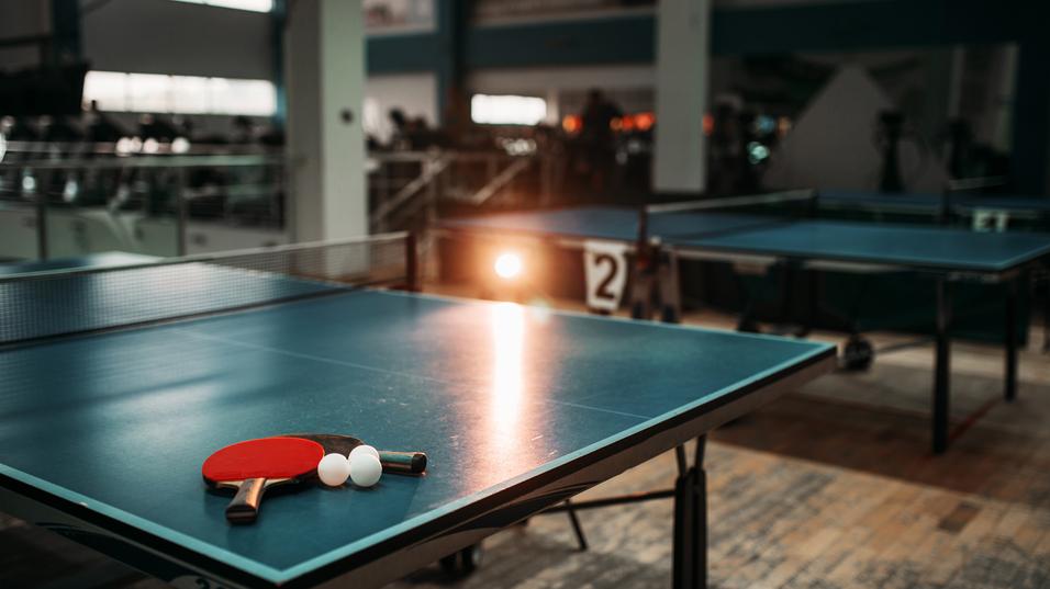 Tischtennistisch mit zwei Schlägern und drei Bällen in einer Sporthalle, im Hintergrund weitere Tischtennistische sichtbar.