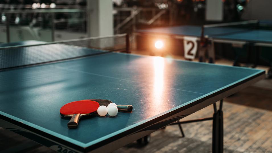 Tischtennistisch mit zwei Schlägern und drei Bällen in einer Sporthalle, im Hintergrund weitere Tischtennistische sichtbar.