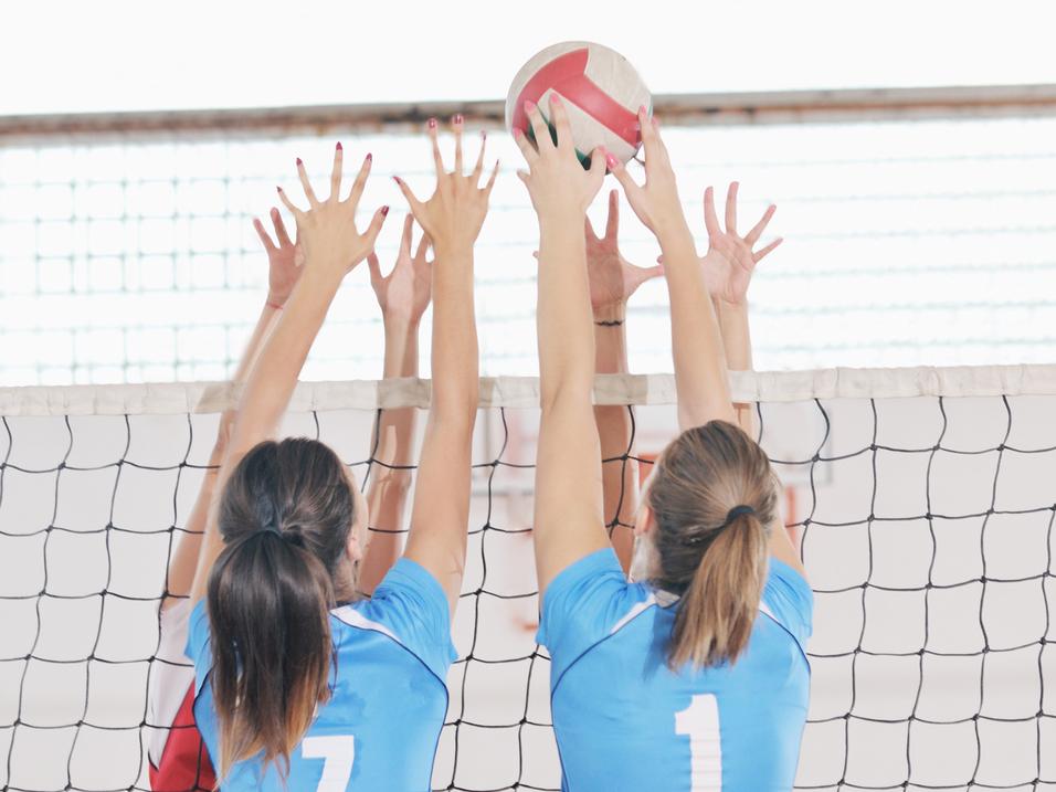 Zwei Volleyballspielerinnen springen am Netz, um einen Ball abzuwehren, während ihre Hände in der Luft sind.