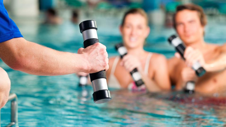 Hand eines Trainers mit Hanteln, während zwei Schwimmer im Wasser mit Hanteln trainieren. Schwimmbecken im Hintergrund.