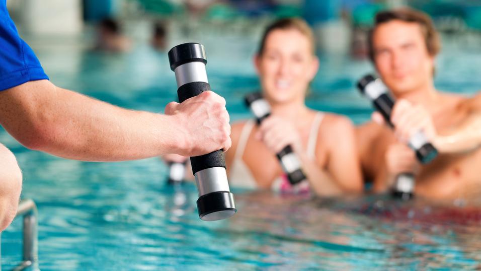Hand eines Trainers mit Hanteln, während zwei Schwimmer im Wasser mit Hanteln trainieren. Schwimmbecken im Hintergrund.