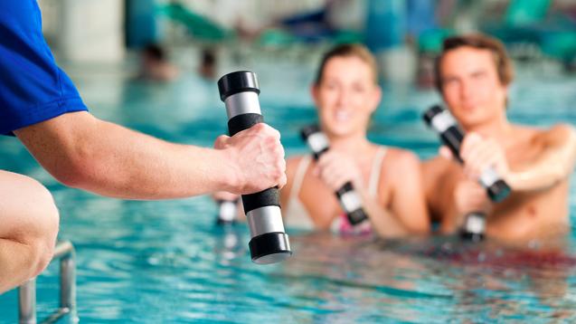 Hand eines Trainers mit Hanteln, während zwei Schwimmer im Wasser mit Hanteln trainieren. Schwimmbecken im Hintergrund.